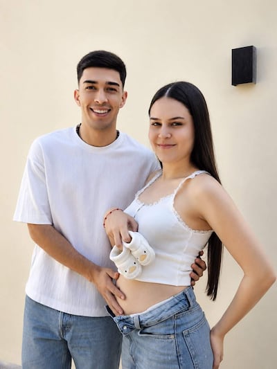 Hombre sonriente viste camiseta blanca y jeans, mientras mujer sostiene zapatos de bebé y sonreír en fondo neutral.