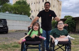 Hombre de camiseta negra asiste a dos niños en sillas de ruedas; mujer sonriente a la izquierda, ambos niños expresan felicidad.