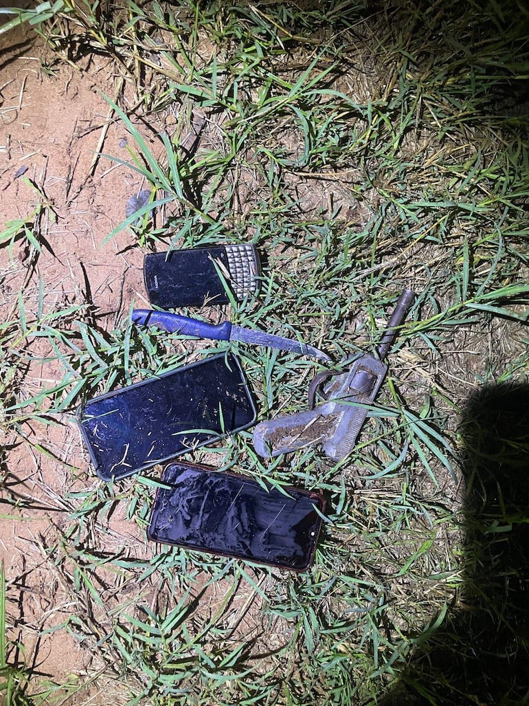 Variados objetos sobre pasto, incluyendo teléfonos móviles y un objeto metálico, sin personas visibles en la escena.