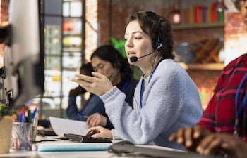 Mujer con auricular gesticula mientras habla, rodeada de dos colegas en un ambiente creativo con paredes de ladrillo expuesto.