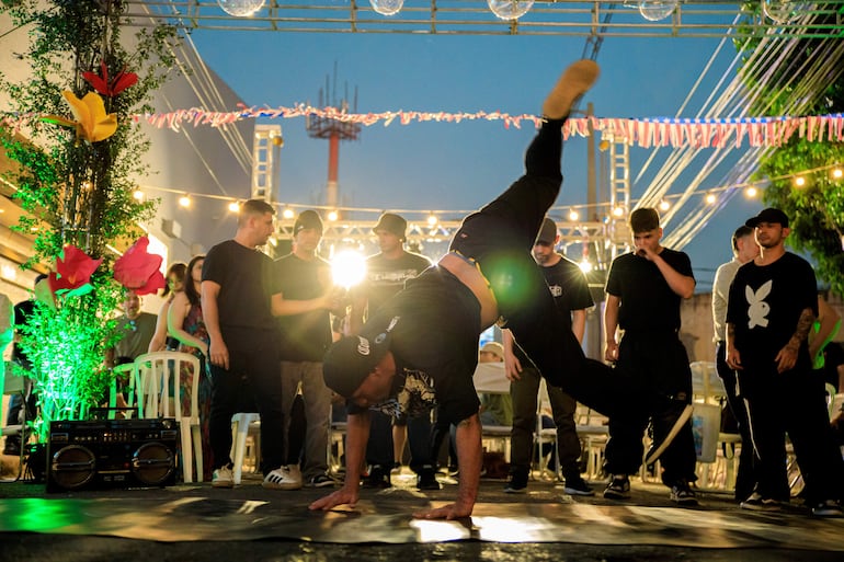 Bailarín realiza acrobacia sobre una mano mientras otros observan, rodeados de luces de colores y música.