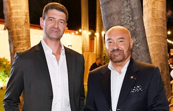 Dos hombres sonrientes, uno con traje oscuro y el otro con blazer, rodeados de vegetación y luces festivas.