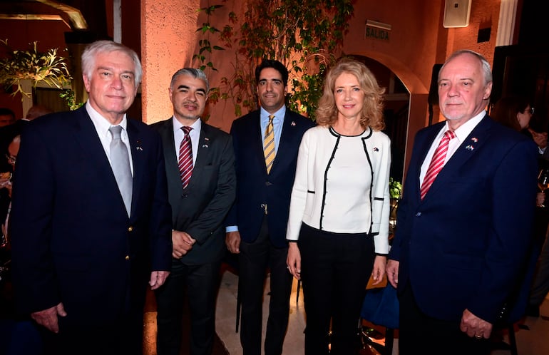 Cinco personas posan en evento: hombre de traje oscuro, hombre con barba y traje gris, hombre con corbata amarilla, mujer con abrigo blanco y hombre con blazer azul y corbata roja.