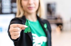 Mujer de cabello rubio y blusa verde sostiene un lazo verde en un evento social, comunicando un mensaje de apoyo.