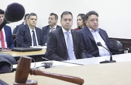 Hombre de traje oscuro y corbata clara en primer plano, rodeado de hombres serios y una mujer atenta en el tribunal.
