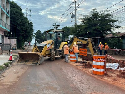Cinco trabajadores con cascos amarillos y chaquetas naranjas observan una retroexcavadora en plena obra pública, rodeados de señalización de advertencia.