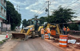 Cinco trabajadores con cascos amarillos y chaquetas naranjas observan una retroexcavadora en plena obra pública, rodeados de señalización de advertencia.