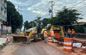 Cinco trabajadores con cascos amarillos y chaquetas naranjas observan una retroexcavadora en plena obra pública, rodeados de señalización de advertencia.