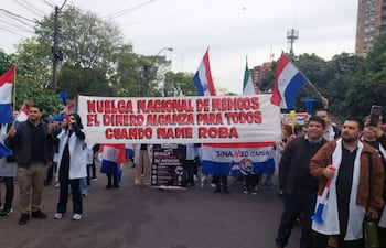 Médicos con batas blancas sosteniendo un cartel que dice 'HUELGA NACIONAL DE MEDICOS' en una calle rodeada de árboles.
