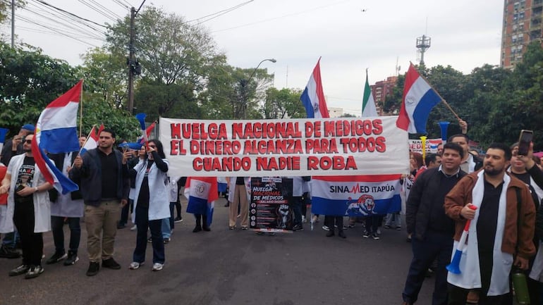 Médicos con batas blancas sosteniendo un cartel que dice 'HUELGA NACIONAL DE MEDICOS' en una calle rodeada de árboles.