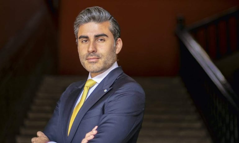 Luis Martín Bogdanovich, con traje oscuro y corbata amarilla, de pie con brazos cruzados, frente a una escalera en fondo marrón.