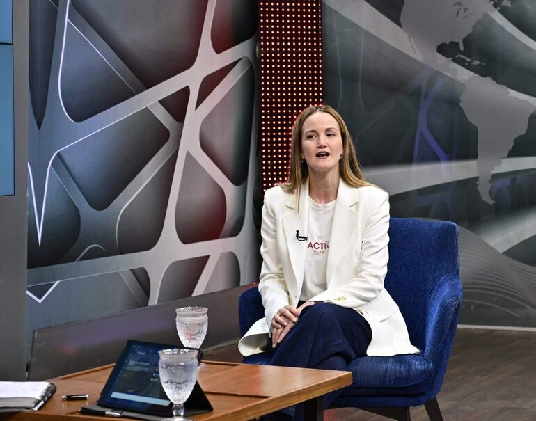 Soledad Núñez sentada en sillón azul, luce chaqueta blanca y camiseta con texto 'ACTIÓN' en un ambiente de estudio moderno.