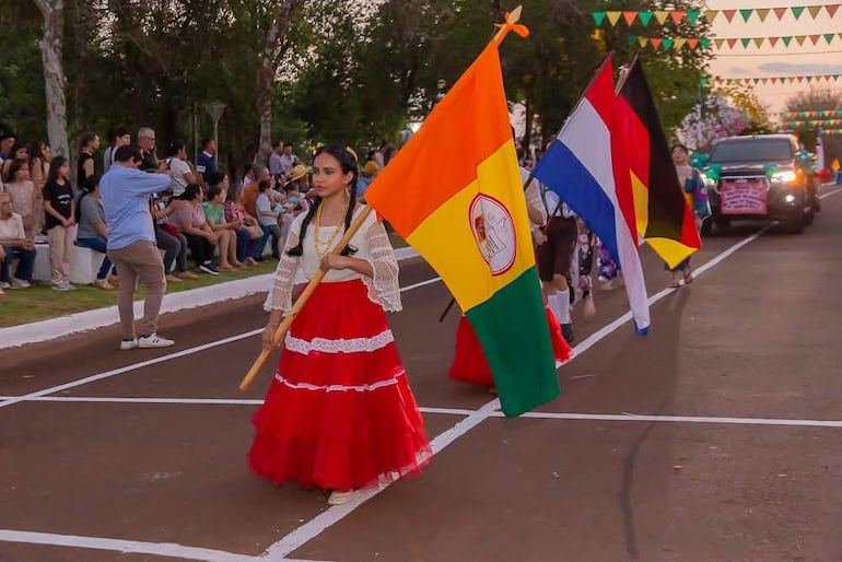 Mujer con vestido rojo y blanco sostiene bandera paraguaya, rodeada de multitudes que observan el desfile festivo.