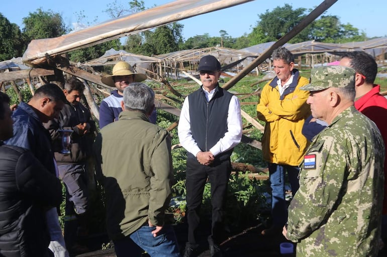 Óscar González con chaleco negro y camisa blanca dialoga con un hombre de sombrero de paja en un entorno agrícola.