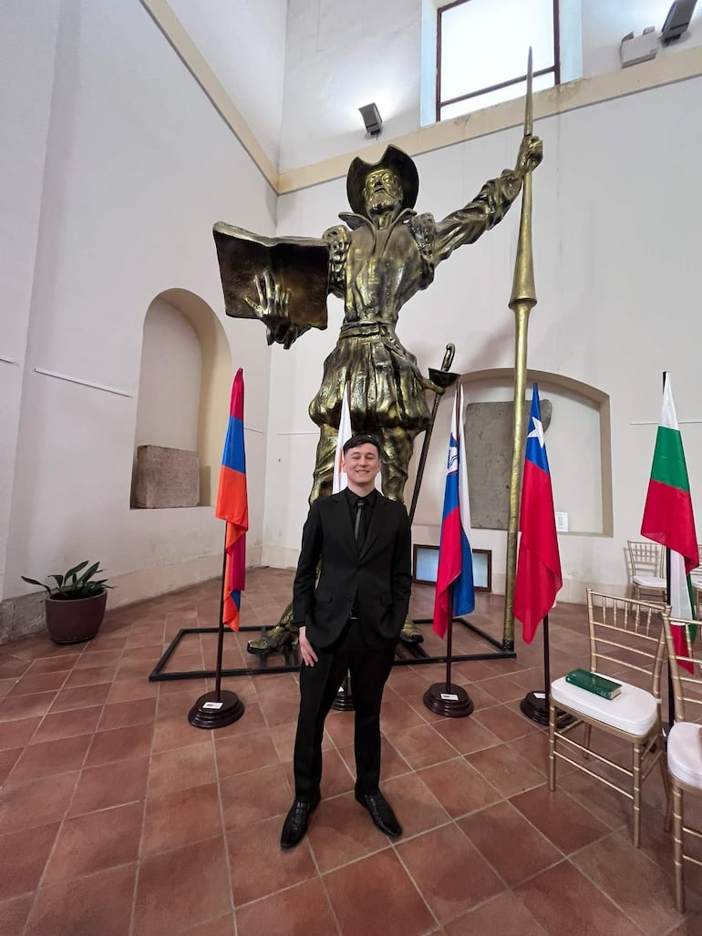 Hombre en traje oscuro junto a escultura dorada y banderas rojas, azules y blancas en un ambiente formal.