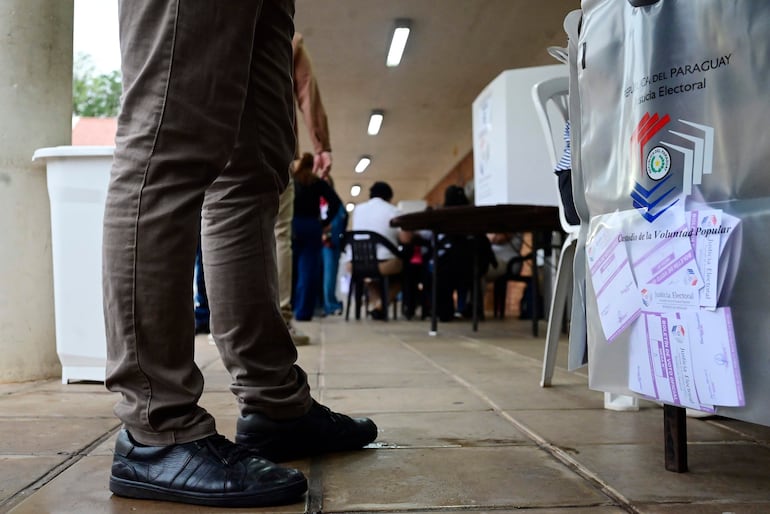 Una persona con pantalones claros y zapatillas negras en una urna electoral con emblema de República del Paraguay.
