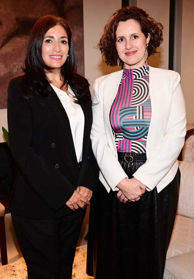 Dos mujeres, una con blusa blanca y otra con patrón colorido, sonríen en un evento formal con ambiente cordial.