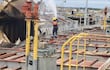 Trabajador con casco y chaleco apagando fuego en barco dañado, con barcos y grúas al fondo en un puerto.