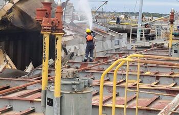 Trabajador con casco y chaleco apagando fuego en barco dañado, con barcos y grúas al fondo en un puerto.