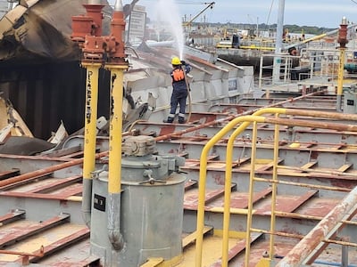 Trabajador con casco y chaleco apagando fuego en barco dañado, con barcos y grúas al fondo en un puerto.