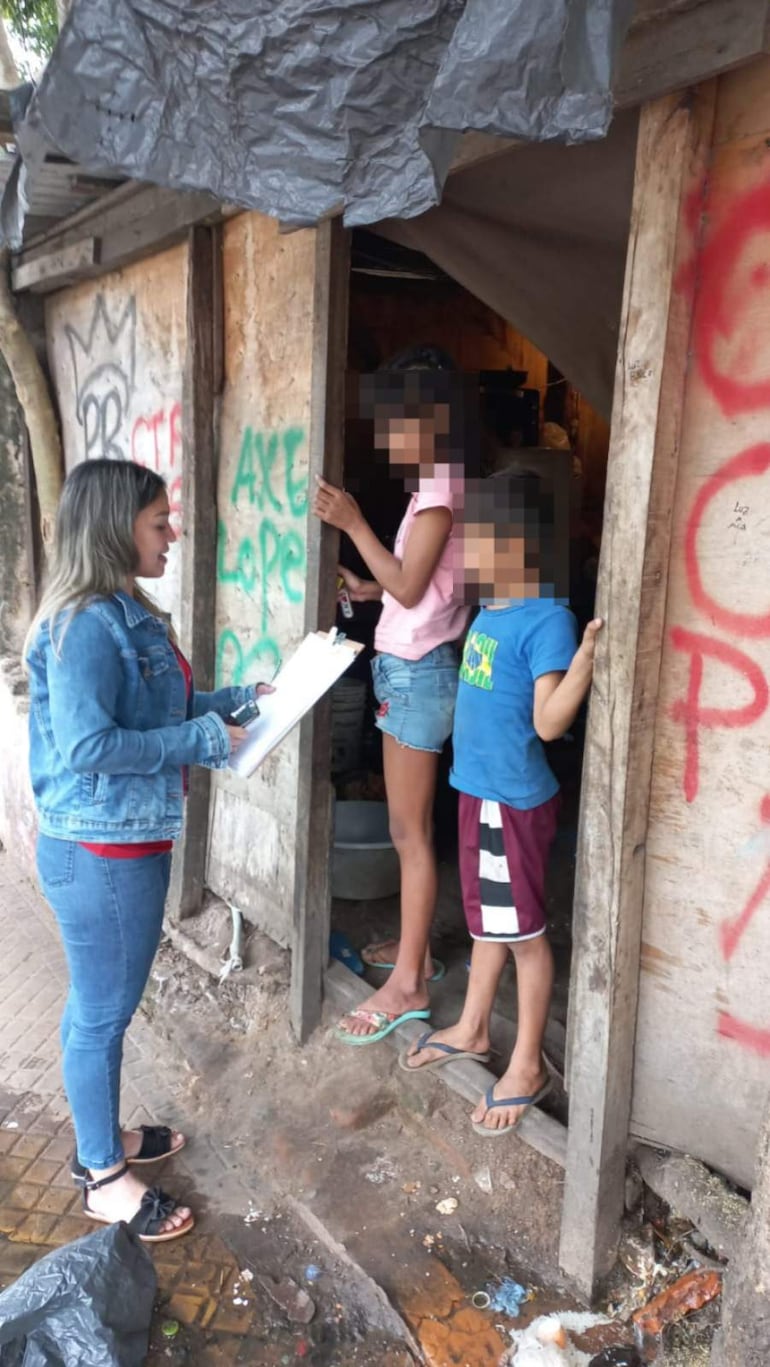 Mujer con chaqueta de jean y blusa roja sostiene un cuaderno, acompañada por dos niños atentos en shorts y camisetas coloridas.