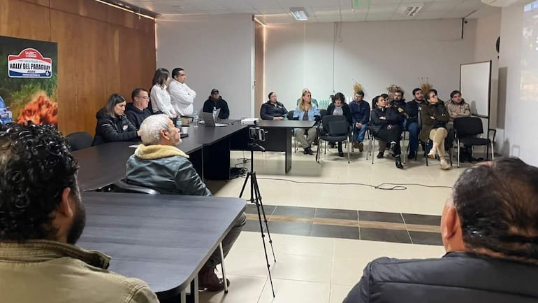 Grupo de 15 personas en sala de conferencias con banner 'RALLY DEL PARAGUAY'. Ambiente formal y atención en presentador.