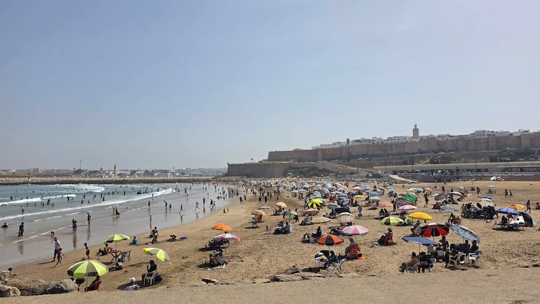 Bañistas en la playa de Rabat, algunos en sombrillas de colores, surfistas en el agua, y al fondo una fortaleza histórica.