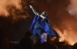 Tan Biónica en escenario con abrigo azul y vestimenta negra, extendiendo un brazo hacia el público rodeado de luces coloridas y humo.