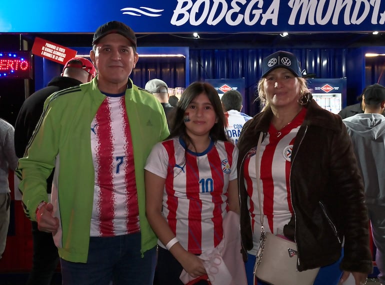 Hombre en camiseta número 7 y chaqueta verde, niña con camiseta número 10, y mujer con escudo de selección en un ambiente festivo.