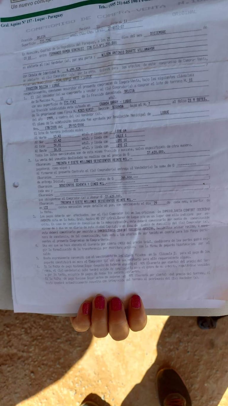 Una mano femenina sostiene un documento de compra-venta en un terreno exterior desprovisto de edificaciones.