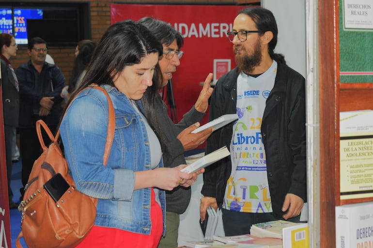 Mujer con chaqueta de mezclilla y pantalones rojos revisa un libro junto a un hombre con barba y camiseta gráfica en evento cultural.