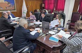 Isaías Ricardo Fretes habla en mesa rodeada por Hassel Jimmy Jiménez y Bettina Albertini, todos revisando documentos en sala con decoración roja.