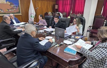 Isaías Ricardo Fretes habla en mesa rodeada por Hassel Jimmy Jiménez y Bettina Albertini, todos revisando documentos en sala con decoración roja.