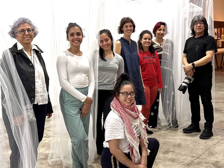 Ocho personas, mayormente mujeres, en ambiente artístico; sonrisas, bufandas y abrigos en un fondo blanco de tela.