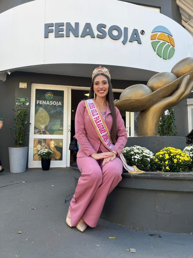 Mujer con corona y vestido rosa sonríe en evento FENASOJA, rodeada de plantas y un monumento agrícola en fondo urbano.