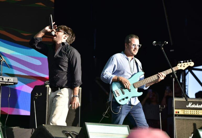 Dos músicos en escenario, uno con micrófono y camisa negra, el otro tocando un bajo azul y camisa clara.
