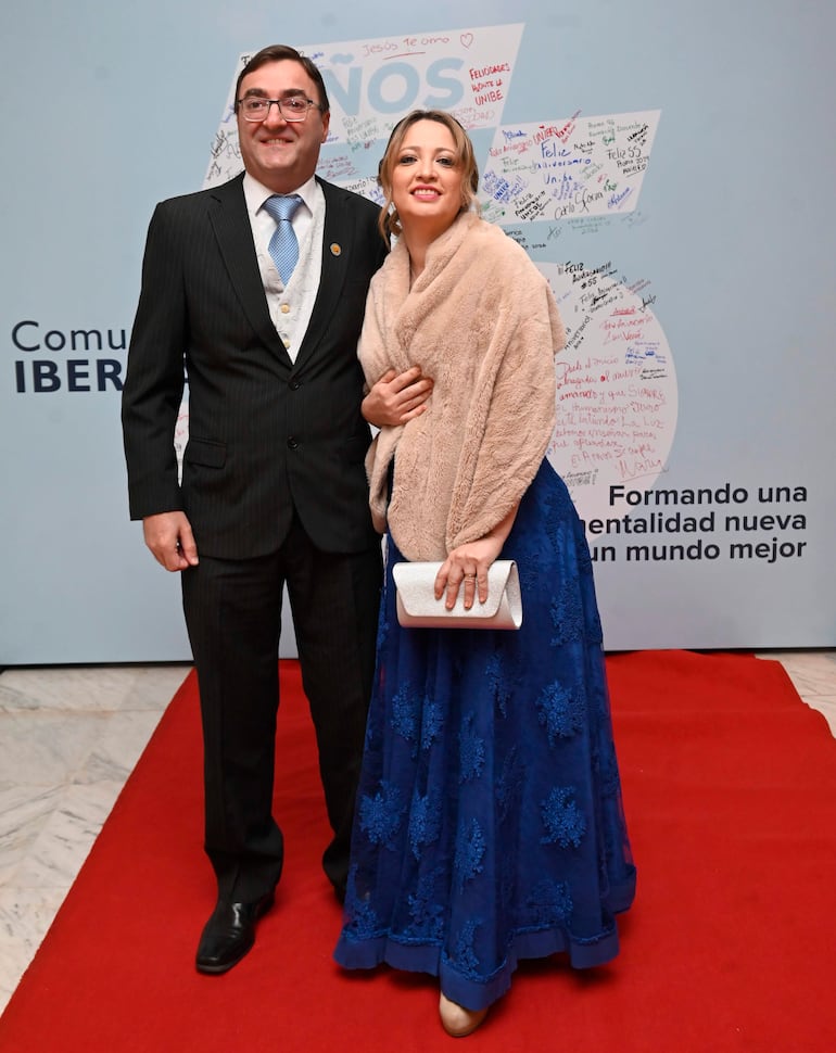 Hombre con traje oscuro y corbata clara, y mujer con vestido azul y abrigo claro, sonríen en elegante evento sobre alfombra roja.