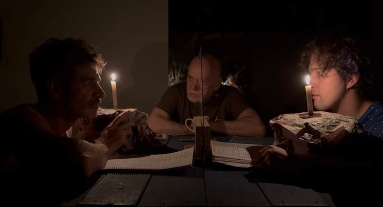 Tres hombres sentados alrededor de una mesa iluminada por velas, con expresiones serias y cuadernos sobre la mesa.