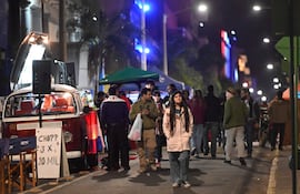Mujer con abrigo claro y hombre en camiseta púrpura interactúan en un evento nocturno lleno de luces y decoración paraguaya.