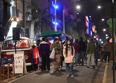 Mujer con abrigo claro y hombre en camiseta púrpura interactúan en un evento nocturno lleno de luces y decoración paraguaya.