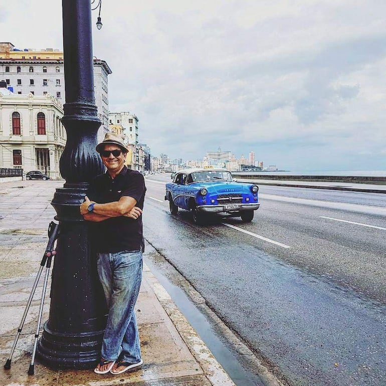 Hombre con camiseta negra y jeans claros sonriendo junto a un coche azul clásico en una calle con edificios y cielo nublado.