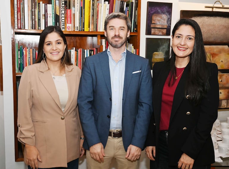 Hombre con barba y cabello gris en blazer azul, flanqueado por dos mujeres de chaquetas claras y oscuras, en un ambiente con estanterías de libros.