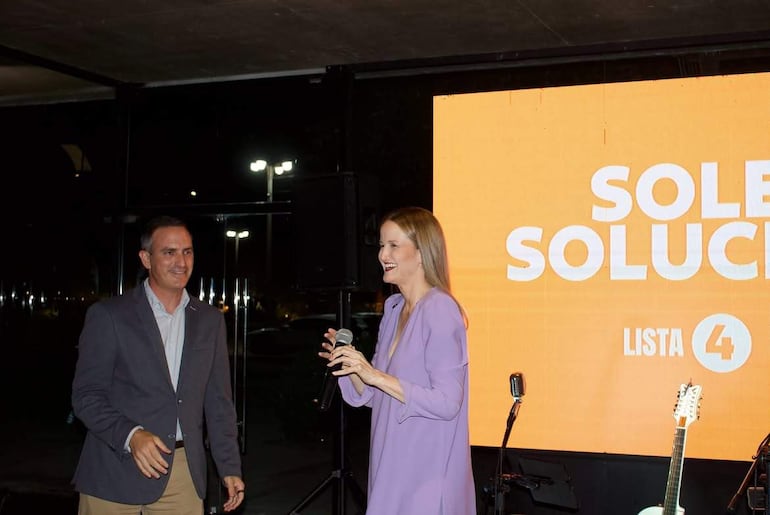 Soledad Núñez en vestido morado y micrófono, sonriente, junto a Stephan Rasmussen en chaqueta gris, ambos en evento con fondo naranja.