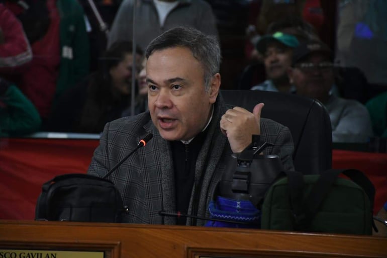 Humberto Blasco sentado en un escritorio con micrófono, gesticulando enfáticamente, mientras una multitud lo escucha en un auditorio.
