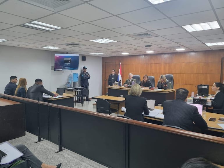 Dos mujeres en el estrado y varios hombres en la audiencia, todos en un ambiente de tribunal formal con paneles de madera.