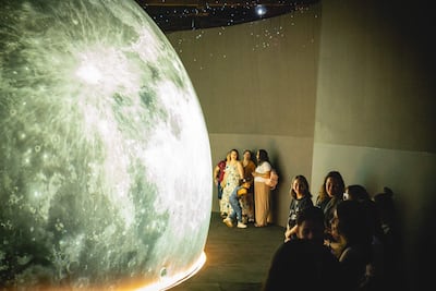 Grupo de mujeres sonrientes junto a una gran representación luminosa de la Luna en ambiente de celebración.