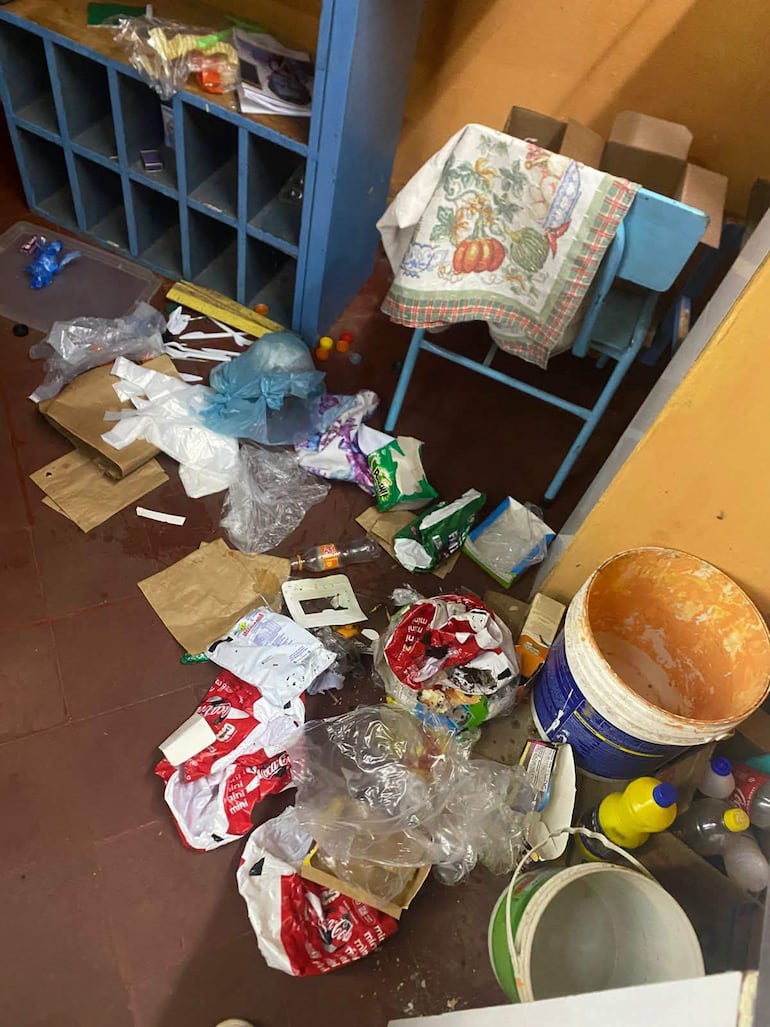 Interior desordenado de un aula, con basura en el suelo y una mesa azul vacía, sin personas presentes.