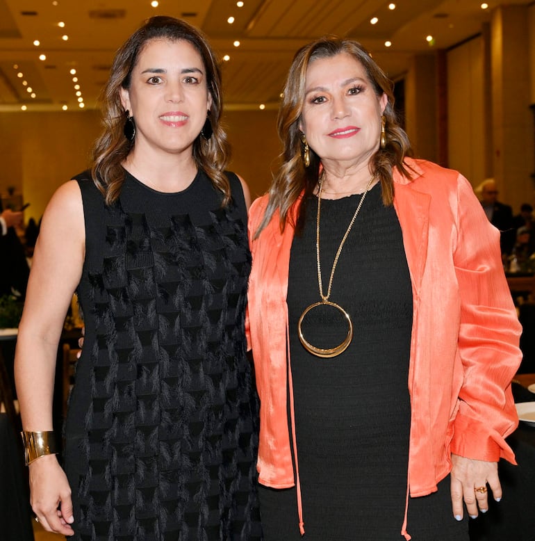 Dos mujeres sonrientes en un salón de eventos, una con vestido negro y otra con blusa naranja, posando juntas para la foto.