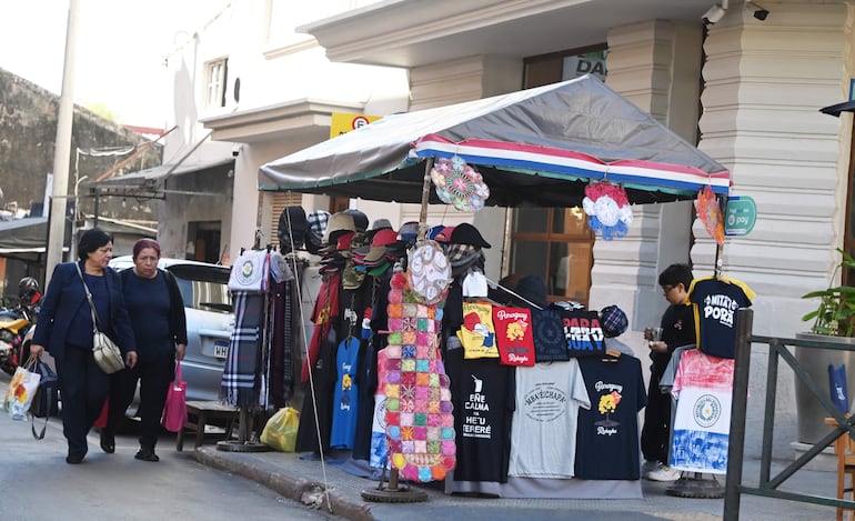 Dos mujeres con ropa oscura y bolsas de compras caminan frente a un kiosco lleno de camisetas y sombreros coloridos.