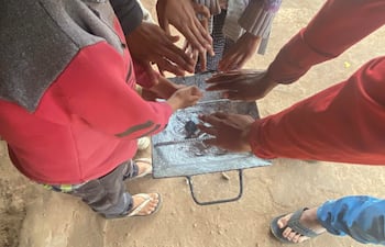 Seis manos de niños y adolescentes rodean una parrilla metálica con carbón encendido, en un entorno de tierra.